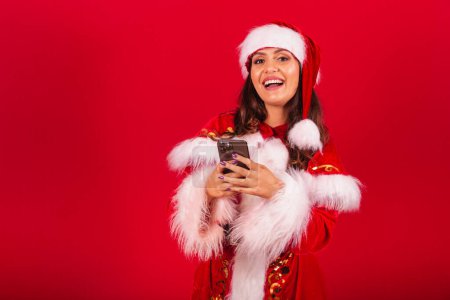Photo for Brazilian woman dressed in Christmas clothes, Santa Claus. holding smartphone and typing. - Royalty Free Image