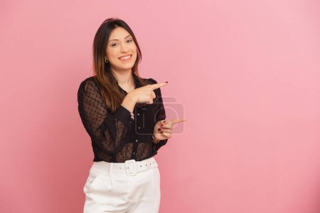 Photo for Beautiful Brazilian Caucasian woman, pink background, smiling, receptive, pointing and showing with finger, a product or advertisement beside her. ad, negative space. - Royalty Free Image