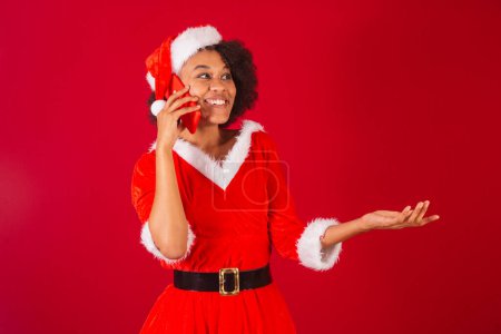 Téléchargez les photos : Belle femme brésilienne noire, habillée en Père Noël claus, maman claus, parlant sur appel vocal par smartphone - en image libre de droit
