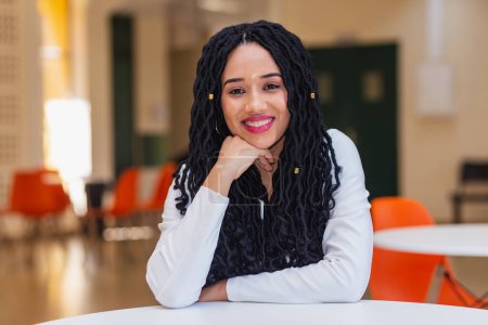 Téléchargez les photos : Jeune femme noire, brésilienne, étudiante dans un couloir universitaire souriante, vêtue de blanc. assis souriant regardant la caméra. - en image libre de droit