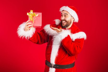 Photo for Brazilian man, dressed in santa claus clothes, giving red present to camera. - Royalty Free Image