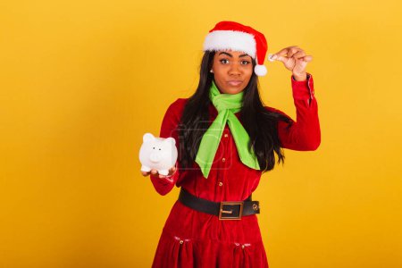 Photo for Beautiful black Brazilian woman, dressed in Christmas clothes, Santa Claus, holding piggy bank and coin. - Royalty Free Image