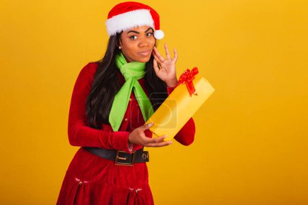 Photo for Beautiful black Brazilian woman dressed in christmas clothes, santa claus, pointing at yellow gift, doubt, - Royalty Free Image