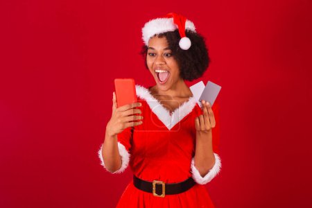 Photo for Beautiful black brazilian woman, dressed as santa claus, mama claus, holding smartphone and credit cards - Royalty Free Image