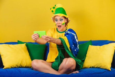 Foto de Mujer caucásica, pelirroja, fan del fútbol brasileño, brasileña, en el sofá viendo el juego por teléfono inteligente - Imagen libre de derechos