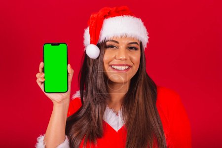 Téléchargez les photos : Belle Brésilienne vêtue de vêtements de Noël, le Père Noël. tenant smartphone montrant écran chromé vert. - en image libre de droit