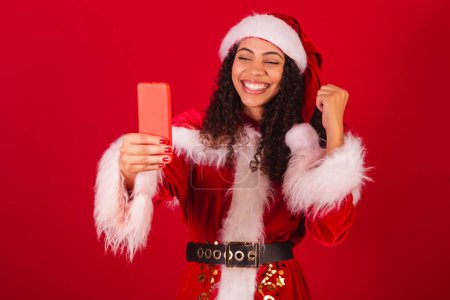 Foto de Hermosa mujer negra brasileña, vestida como santa claus, ropa de Navidad. celebrando con algo en el smartphone. - Imagen libre de derechos