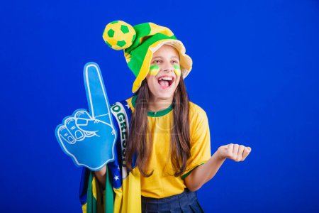 Téléchargez les photos : Jeune fille, fan de football du Brésil. habillé en chapeau et drapeau. en utilisant doigt de mousse, faire la fête. incroyable. - en image libre de droit