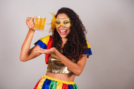 Téléchargez les photos : Belle femme brésilienne noire, avec des vêtements frevo, carnaval. dans le mascara, montrant un verre de bière, faire la fête dans le bar. - en image libre de droit