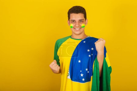 Photo for Young Brazilian man soccer fan. dressed in green, hand on chest, celebrating with the flag of Brazil. - Royalty Free Image