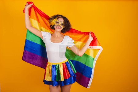 Photo for Beautiful Brazilian woman, dressed in carnival clothes, colorful skirt and white shirt. wearing mascara, holding lgbt flag. - Royalty Free Image