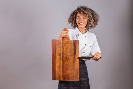 Téléchargez les photos : Jeune Brésilienne noire, cuisinière, chef cuisinière, portant l'uniforme de restaurant. tenant une planche et un couteau en bois pour les textes et les publicités, présentant. - en image libre de droit