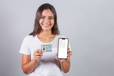 Photo for Brazilian woman, with driver's license, CNH, document. Smartphone, white screen for advertisements. - Royalty Free Image