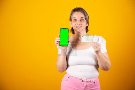 Photo for Adult Brazilian woman holding voter registration card and smartphone with green screen - Royalty Free Image