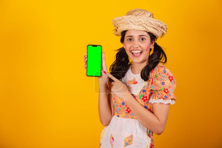 Photo for Beautiful Brazilian woman, with festa junina clothes, holding smartphone with green screen. - Royalty Free Image
