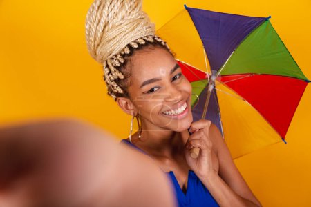 Photo for Beautiful black Brazilian woman, dressed in carnival clothes, blue, and yellow background, bun and braids, frevo umbrella. taking self portrait. - Royalty Free Image