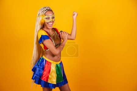 Téléchargez les photos : Belle femme brésilienne noire, vêtue de vêtements de carnaval, masque. fond jaune. tenant des biceps, montrant de la force. - en image libre de droit
