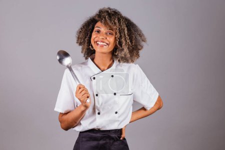 Téléchargez les photos : Jeune Brésilienne noire, cuisinière, chef cuisinière, portant l'uniforme de restaurant. tenant une grande cuillère pour préparer les sauces. - en image libre de droit