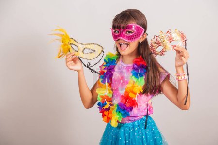 Téléchargez les photos : Belle fille brésilienne, enfant, habillée pour le carnaval au Brésil. avec masque de carnaval, tenant des masques, invitant à la fête. - en image libre de droit