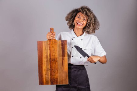 Photo for Young black Brazilian woman, cook, masterchef, wearing restaurant uniform. holding wooden board and knife for texts and advertisements, presenting. - Royalty Free Image