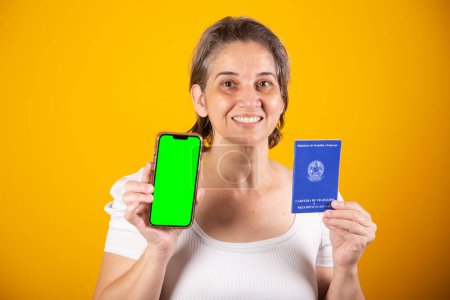 Photo for Adult Brazilian woman, holding work and social security card, smartphone with green screen - Royalty Free Image