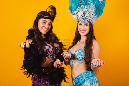 Téléchargez les photos : Deux amies brésiliennes avec des vêtements de reine de l'école de samba, carnaval. Couronne de plumes. invitant avec les mains - en image libre de droit