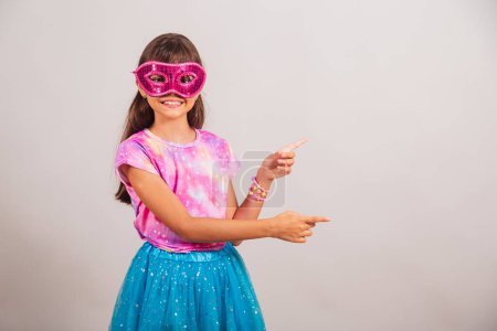 Téléchargez les photos : Belle fille brésilienne, enfant, habillée pour le carnaval au Brésil. avec masque de carnaval, présentant le produit sur le côté. - en image libre de droit