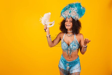 Foto de Mujer negra reina de la escuela brasileña de samba, con ropa de carnaval azul y corona de plumas. sosteniendo máscara. invitando a las manos. - Imagen libre de derechos