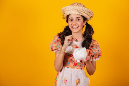 Photo for Beautiful Brazilian woman, with June party clothes, holding coin and piggy bank. - Royalty Free Image