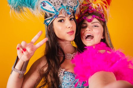 Téléchargez les photos : Deux amies brésiliennes avec des vêtements de reine de l'école de samba, carnaval. Couronne de plumes. prise d'autoportrait avec smartphone. - en image libre de droit