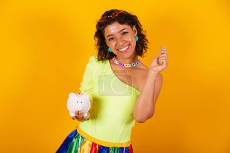 Photo for Beautiful afro american brazilian woman in carnival clothes holding piggy bank and coin. - Royalty Free Image