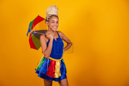 Foto de Hermosa mujer brasileña negra, vestida con ropa de carnaval, fondo azul y amarillo, moño y trenzas, sonriendo con paraguas frevo. - Imagen libre de derechos