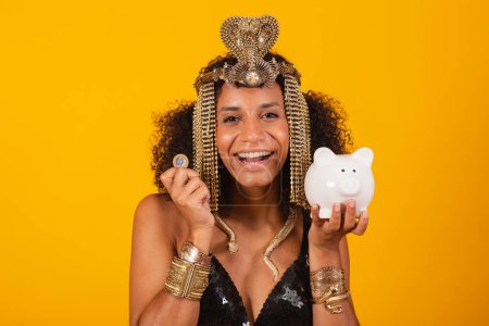 Téléchargez les photos : Belle femme noire brésilienne, en vêtements de carnaval Cleopatra, tenant tirelire et pièce de monnaie. - en image libre de droit