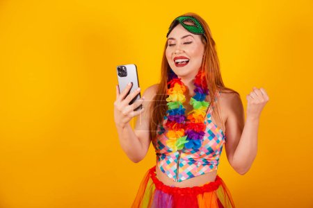 Photo for Brazilian redhead, in carnival clothes, with a flower necklace around her neck, celebrating with a smartphone. - Royalty Free Image
