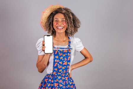 Foto de Joven brasileña negra, con ropa de campo, vestida para Festa Junina. celebración de teléfono inteligente con pantalla blanca para anuncios, aplicaciones móviles. - Imagen libre de derechos