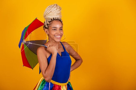 Téléchargez les photos : Belle femme brésilienne noire, vêtue de vêtements de carnaval, fond bleu et jaune, chignon et tresses, souriant avec parapluie frevo. - en image libre de droit