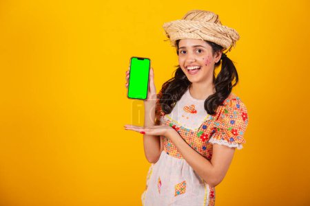 Téléchargez les photos : Belle femme brésilienne, avec des vêtements Festa junina, tenant smartphone avec écran vert. - en image libre de droit