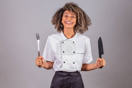Photo for Young black Brazilian woman, cook, masterchef, wearing restaurant uniform. holding fork and knife for barbecue. Meat cutting. - Royalty Free Image