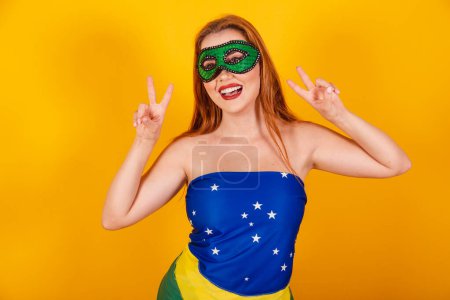 Photo for Beautiful brazilian redhead girl with carnival clothes made with flag of brazil, making peace and love sign. - Royalty Free Image