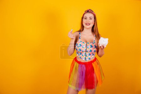Photo for Brazilian redhead, in carnival clothes, holding piggy bank and coin. - Royalty Free Image