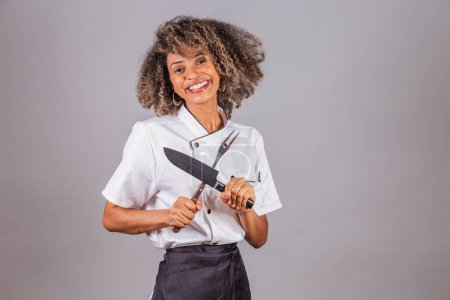 Téléchargez les photos : Jeune Brésilienne noire, cuisinière, chef cuisinière, portant l'uniforme de restaurant. fourchette et couteau pour barbecue. Découpe de viande. - en image libre de droit