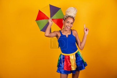 Foto de Hermosa mujer brasileña negra, vestida con ropa de carnaval, fondo azul y amarillo, moño y trenzas, sonriendo con paraguas frevo. bailando - Imagen libre de derechos