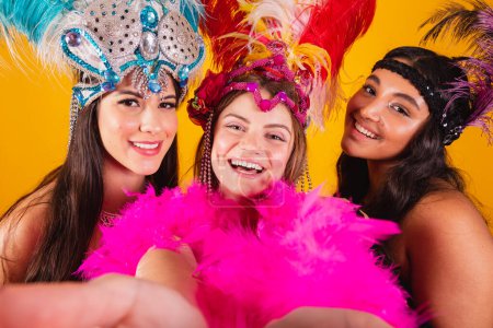 Foto de Tres amigas brasileñas con ropa de reina de la escuela de samba, carnaval. Corona de plumas. tomando autorretrato con smartphone. - Imagen libre de derechos