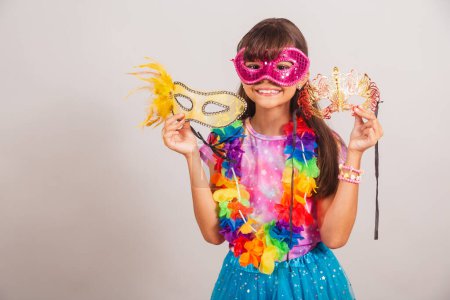 Téléchargez les photos : Belle fille brésilienne, enfant, habillée pour le carnaval au Brésil. avec masque de carnaval, tenant des masques, invitant à la fête. - en image libre de droit
