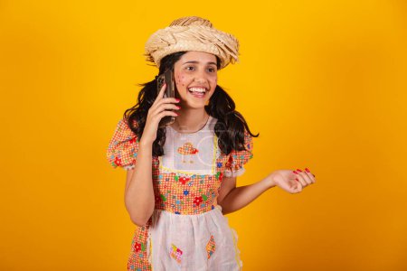 Téléchargez les photos : Belle femme brésilienne, avec des vêtements de fête de juin, parlant sur appel vocal par smartphone. - en image libre de droit