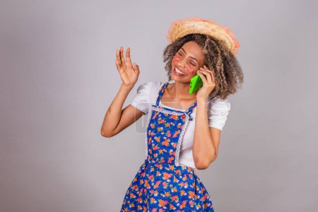 Photo for Young black Brazilian woman, with country clothes, dressed for Festa Junina. Saint John's festival. on voice call with smartphone. - Royalty Free Image