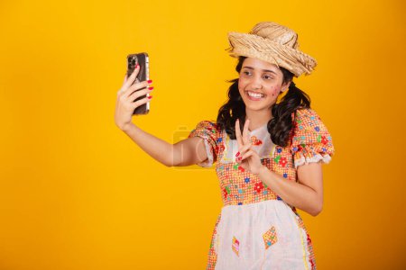 Téléchargez les photos : Belle femme brésilienne, avec des vêtements de festa junina, prenant autoportrait avec smartphone. - en image libre de droit