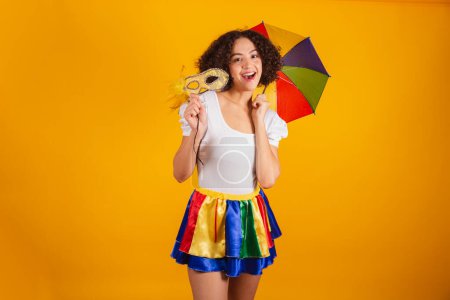 Téléchargez les photos : Belle femme brésilienne, vêtue de vêtements de carnaval, jupe colorée et chemise blanche. parapluie frevo, tenant masque de carnaval. - en image libre de droit