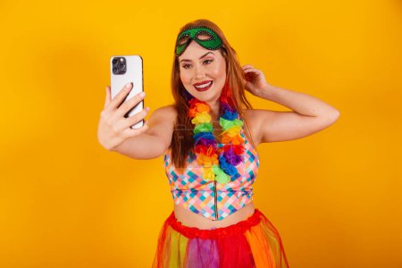 Foto de Pelirroja brasileña, vestida de carnaval, con un collar de flores alrededor del cuello, tomando una selfie con un smartphone. - Imagen libre de derechos