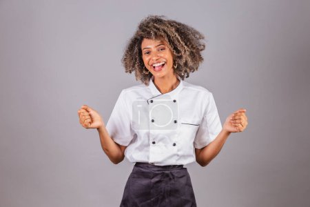 Téléchargez les photos : Jeune Brésilienne noire, cuisinière, chef cuisinière, portant l'uniforme de restaurant. célébrant, extrêmement heureux. - en image libre de droit